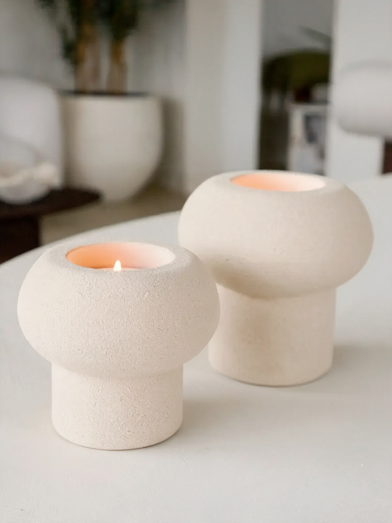 Twee beige aardewerk waxinelichthouders op voet met brandend waxinelicht in Japandi interieur – STILL